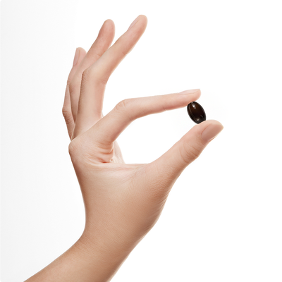 Hand holding a small dark brown oval supplement capsule between index finger and thumb