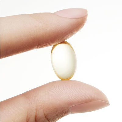 Hand holding a single golden yellow oval fish oil softgel capsule between two fingers