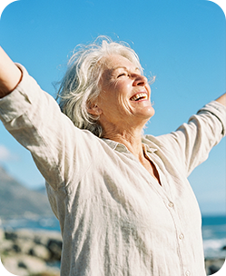 Elderly woman with white hair raising arms joyfully outdoors near ocean on sunny day