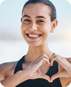 Young woman in black top making heart shape with hands, smiling outdoors near water