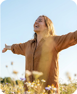 Woman in brown linen shirt, arms outstretched, laughing joyfully in a sunny wildflower field