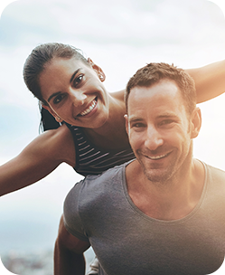 Smiling couple outdoors, woman on man's back, both laughing in bright light