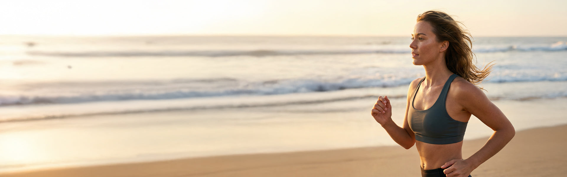 Woman in gray sports bra running on sandy beach at golden sunset with ocean waves behind her