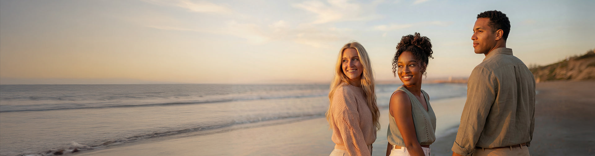Three friends smiling on a sandy beach at golden sunset, ocean waves behind them